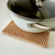Saucepan resting on a square bamboo laser-cut trivet with geometric grooves on a countertop.