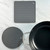 Top-down view of a square gray silicone hot pad with hanging eyelet, a round honeycomb trivet, and a black skillet on a light countertop