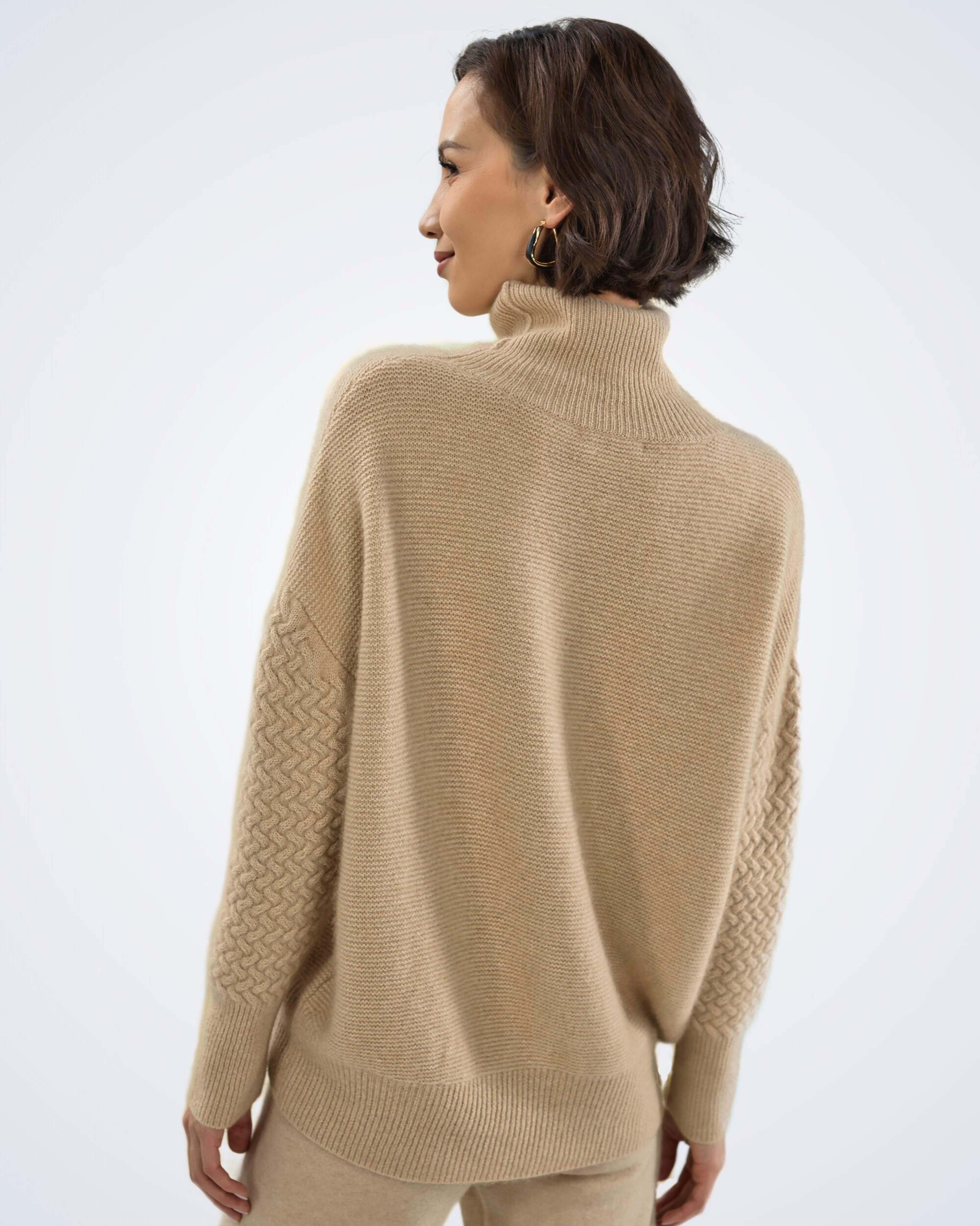 Unur model wearing an oversized natural beige cashmere jumper with cable-knit sleeves and a roll neck, rear view