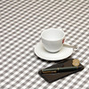 Acrylic Oilcloth - Mirha Yarn Dyed: Gingham Check - Grey. Pictured with a cup and saucer, phone, and pen.