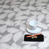 Wipe clean tablecloth. Living Feathers - Grey. Pictured with a cup and saucer, 'phone, pen and coins.