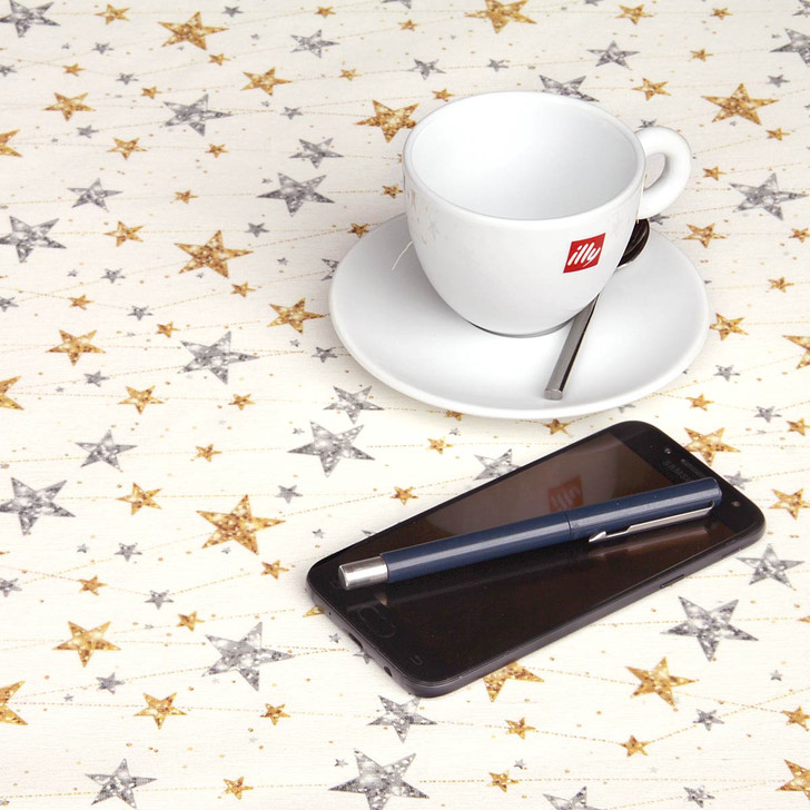 Navarra Silver & Gold Stars Wipe Clean Tablecloth. Pictured on a table with a phone, cup and pen