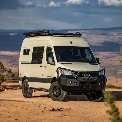 Beige Mercedes Sprinter van outfitted with several off-road accessories, parked near open canyons featuring Aluminess Explorer Roof Rack.