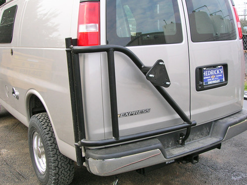 A tan-colored Chevy Express van with a driver side rear door tire rack from Aluminess Products.