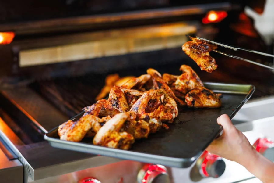 Grilled chicken wings being lifted with tongs above a tray in front of an open stainless steel grill