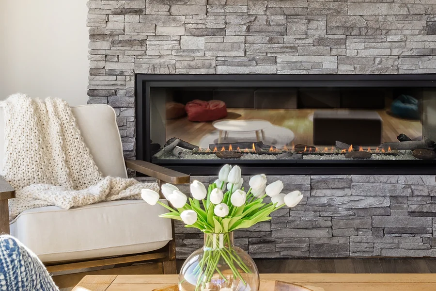 Wide gas fireplace in a gray stone wall beside a cream armchair and a vase of white tulips