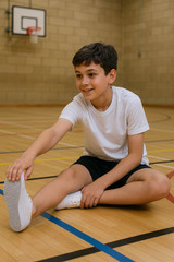 Child wearing white lace-up school plimsolls, stretching in a gymnasium with court markings, dressed in a white T-shirt and black shorts.