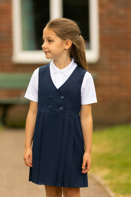 Girl wearing a navy four-button V-neck school pinafore dress with box pleats, layered over a white polo shirt, standing outdoors in a natural school setting, looking away from the camera.
