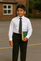 Boy wearing black plus size sturdy fit school trousers, white shirt and black tie, holding school books while standing in a playground