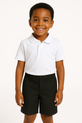 The image features a smiling boy, aged about 7, posed against a plain white backdrop. He is dressed in a neat white polo shirt and black school shorts, radiating confidence and charm. His natural expression and tidy appearance make this a great representation of a school uniform style.