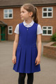 Girls royal blue zip-front box pleat school pinafore dress with heart-shaped zipper pull, styled over a white polo shirt in a natural schoolyard setting – smart, comfortable and durable uniform from School Wear United.