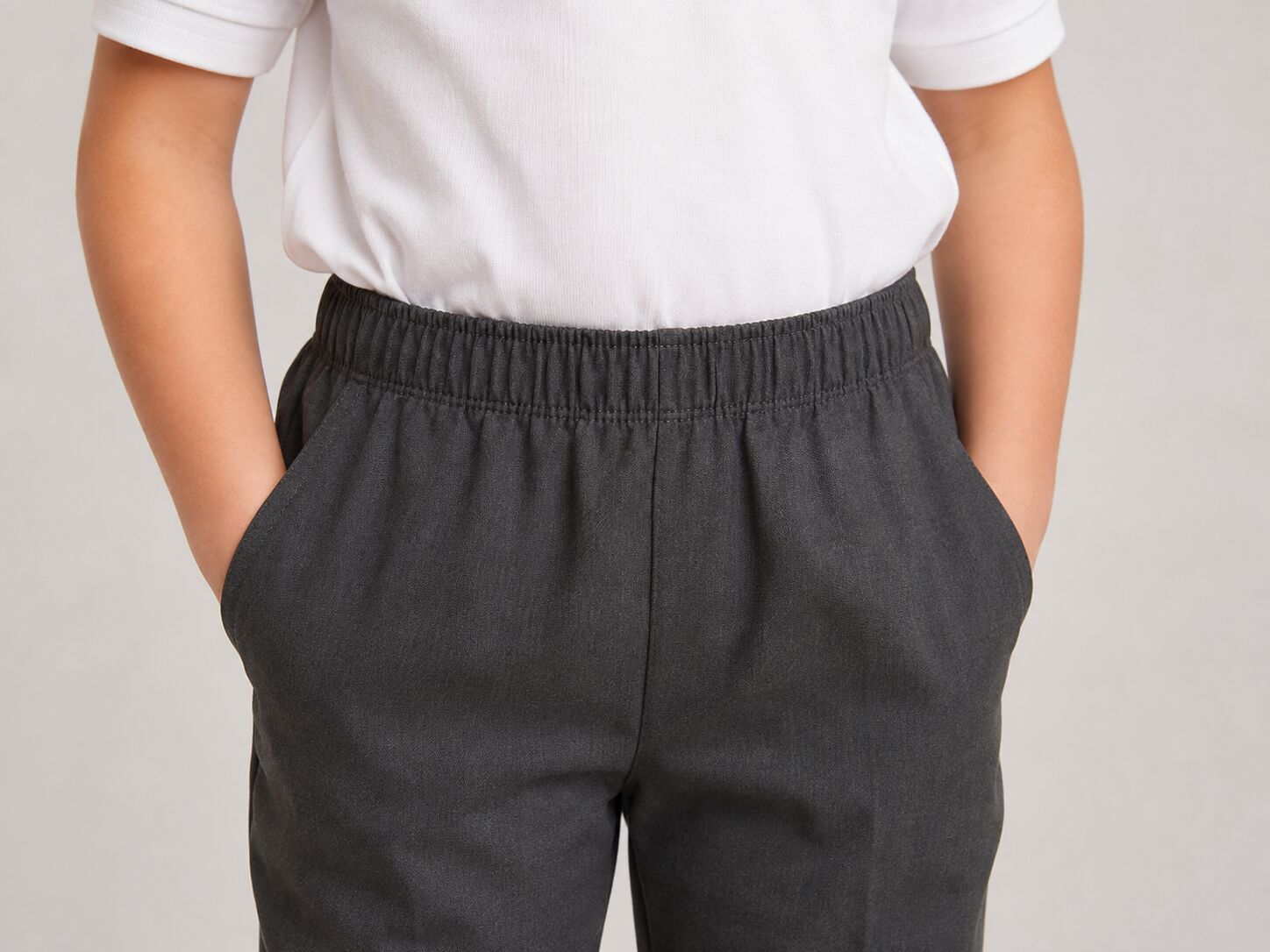 Close up of grey school trousers with a full elastic pull on waistband, no zip and no button, worn by a child with hands in pockets
