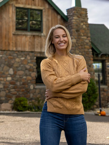 Woman standing in front of a wood cabin during fall wearing the Winter Wisteria Embroidered Cable Knit Sweater in mustard yellow.