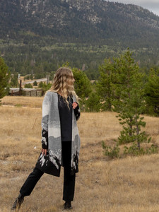 Woman standing in field during fall day wearing the Vista Jacquard Mountain Cardigan Sweater in grey mountain border print.