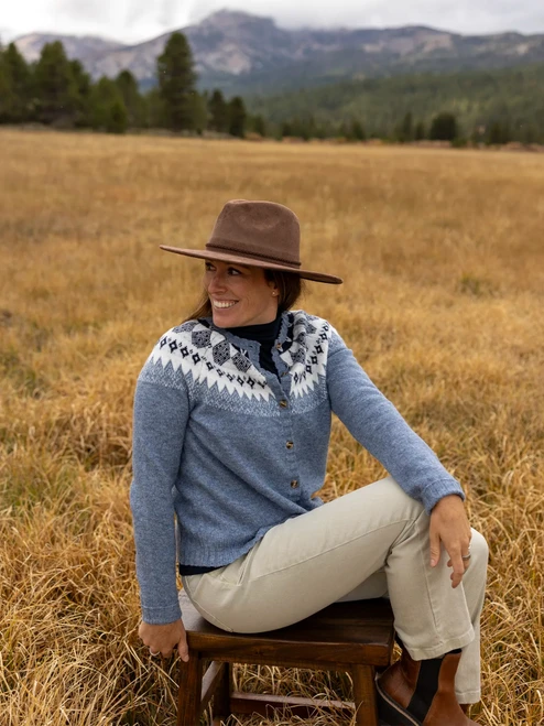 Woman sitting in field during fall wearing the Reine Fair Isle Cardigan Sweater in stonewash blue.