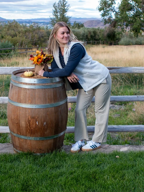 Woman leaning on wine barrel during fall wearing the Alabaster Pine Sweater Knit Fleece Vest in whisper white.
