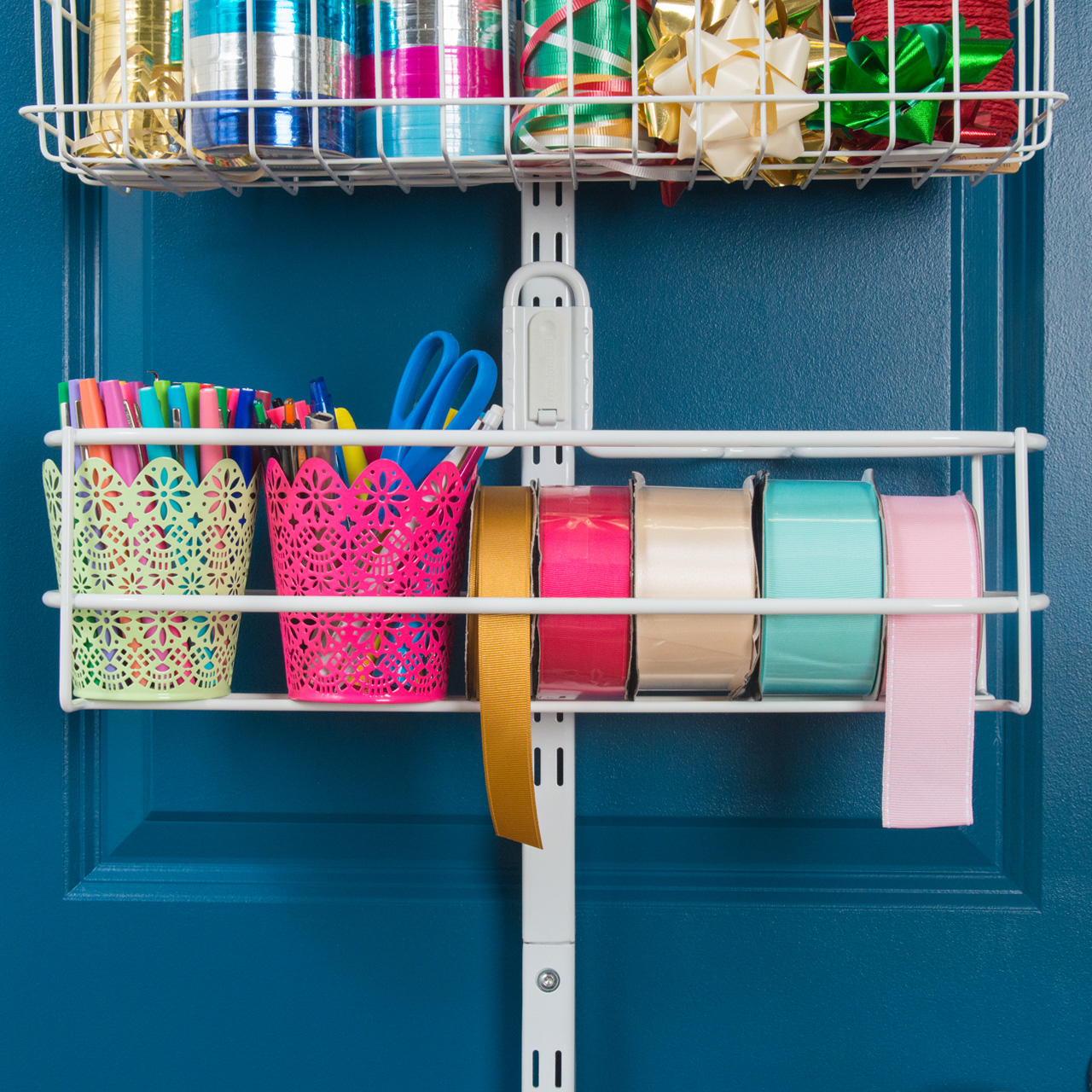 #close up image of a white basket containing ribbons and art supplies such as pens and scissors in a basket. Product is white in color mounted to a blue door