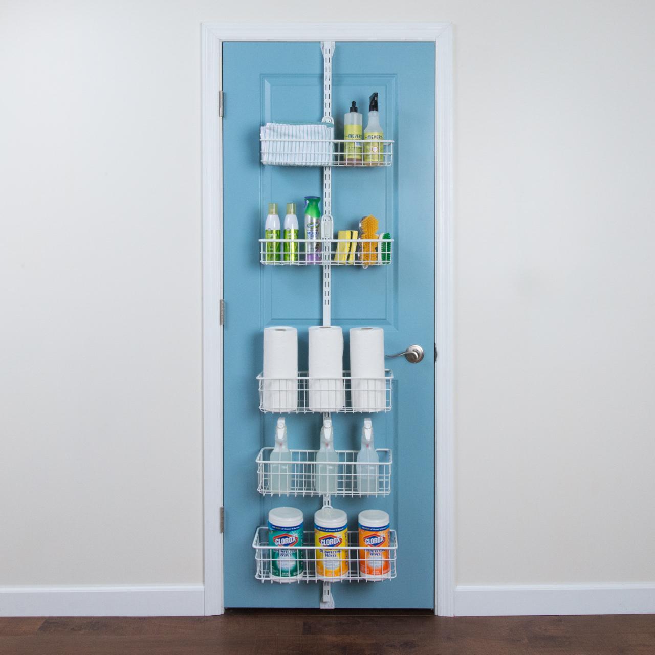 # White Over the Door storage system mounted to a blue door containing various sized baskets with cleaning supplies and paper towels. 