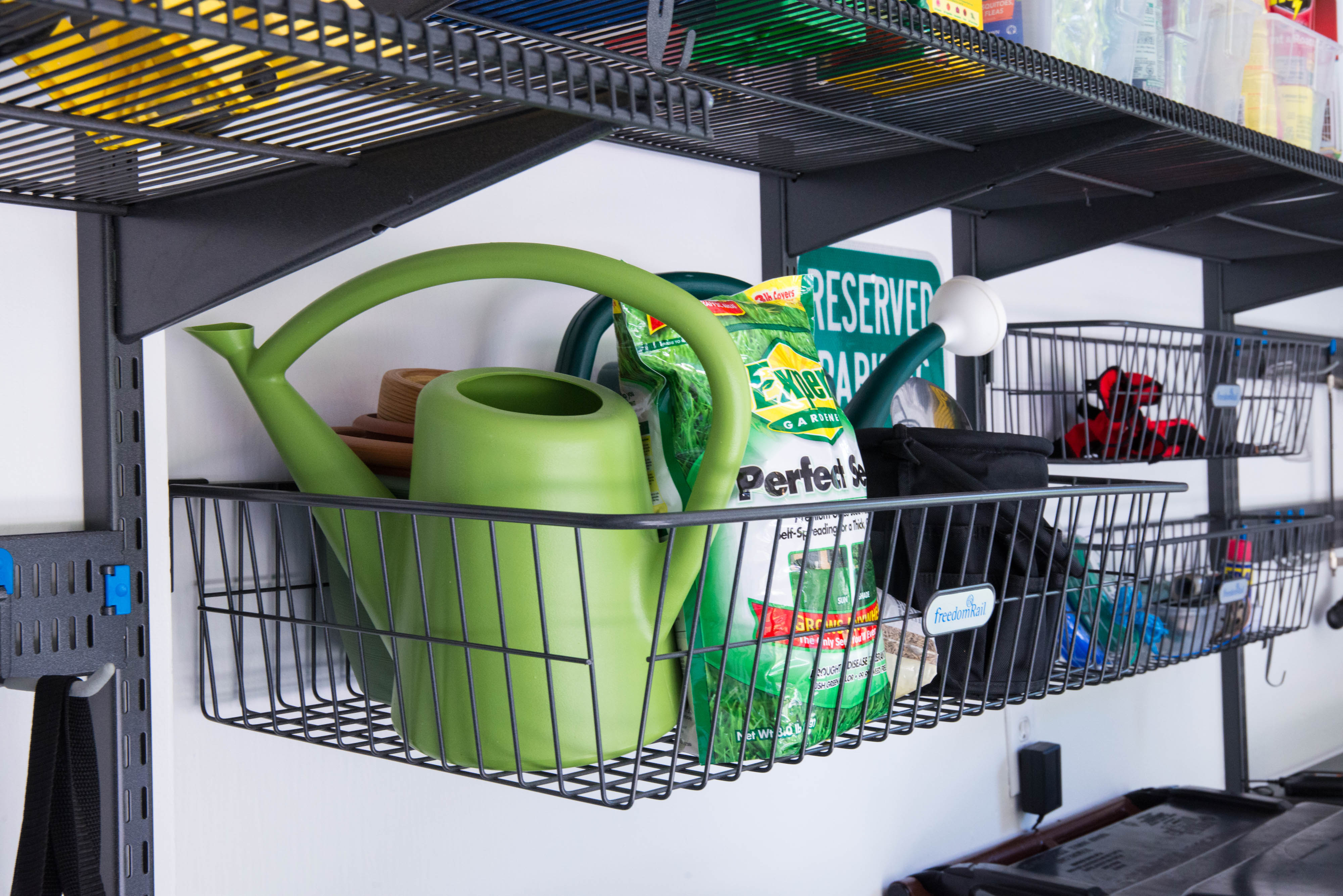 This is an Organized Living freedomRail Garage with granite gray ventilated shelves and baskets. The walls are white and the baskets are holding green watering cans and garden supplies. 