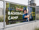 Mesh banner promoting a youth baseball camp, mounted on outdoor fencing where the perforated vinyl lets wind pass through without damaging the banner or its hardware.