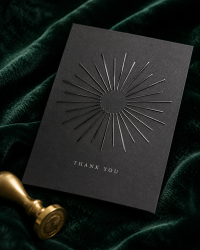 Raised spot UV greeting card in matte charcoal cardstock with a glossy sunburst pattern and "Thank You" lettering, styled with a gold wax seal on emerald velvet.