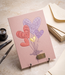 Embossed birthday card in blush pink cardstock with raised heart balloon design in coral and lavender, small gold foil accent tag, styled with a fountain pen and cream envelope.