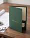 Embossed presentation folder in forest green cardstock with a subtle geometric building mark and gold foil "Studio Proposal" lettering, styled on a desk with rolled blueprints.