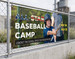 Mesh banner promoting a youth baseball camp, mounted on outdoor fencing where the perforated vinyl lets wind pass through without damaging the banner or its hardware.