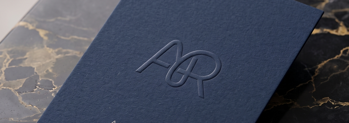 A dark navy business card with a blind embossed monogram ("AR") raised above the surface, no color or foil added, letting the paper's own texture and shadow carry the mark.