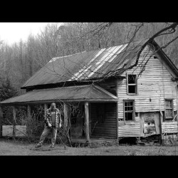 Black and White Image of Greg Pickens of Finding America Metal Detecting In Front of Old Rustic House with Bare Trees Around It and in the Background