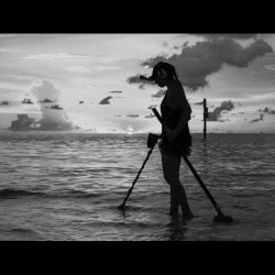 Silhouette of She Detector Water Hunting for Treasures at the beach with Garrett Metal Detector in Front of her and Sand Scoop Behind with Cloudy Sky in Background all in Black and White