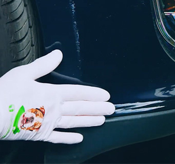 Gloved hand pointing to scratches remaining on a dark blue vehicle bumper after removing paint transfer.