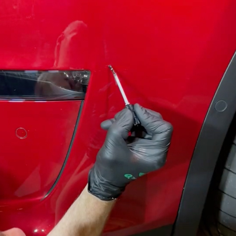 Black-gloved hand applying clearcoat with a fine brush to a red vehicle door.