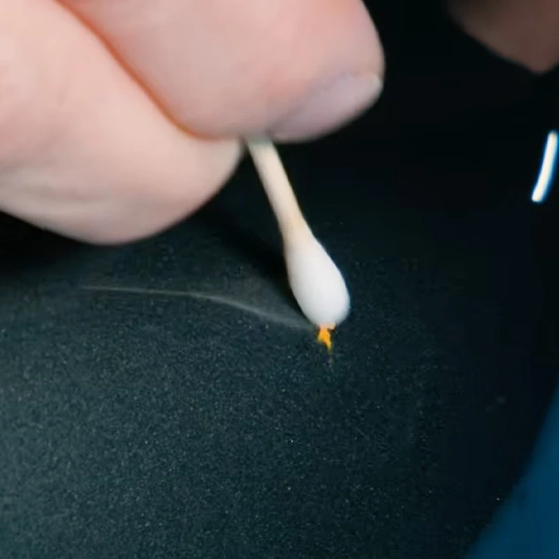 Close-up of a hand using a cotton swab on a small defect in a dark painted surface.