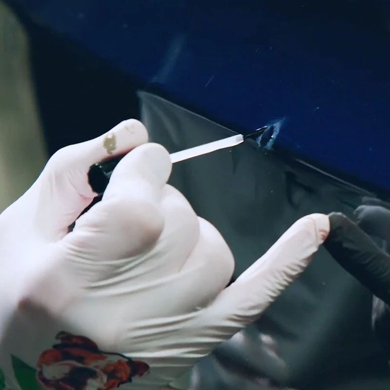 Gloved hand applying primer with a fine brush to a scratch on a dark blue vehicle panel