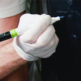Gloved hand applying blue colorcoat with a touch up paint pen to small chips on a dark vehicle panel.