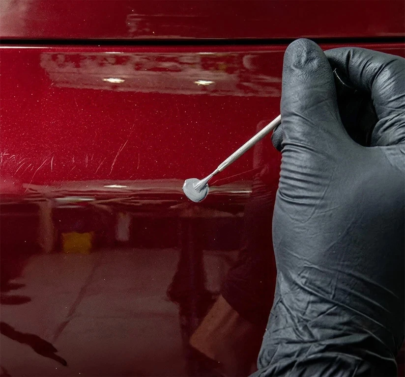 Gloved hand applying a thick layer of gray primer to a red vehicle panel