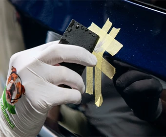 Gloved hand wet sanding a masked primer repair on a dark blue vehicle panel