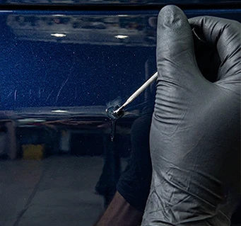 Gloved hand applying colorcoat to a blue vehicle panel without finishing the repair with clearcoat