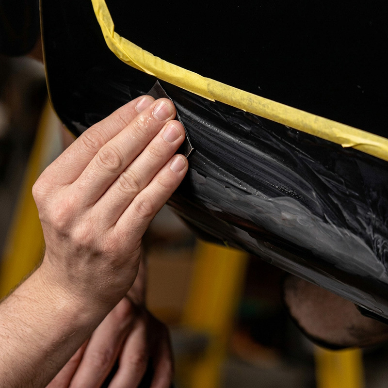 and sanding a large scratch on a black vehicle panel inside a taped repair area.