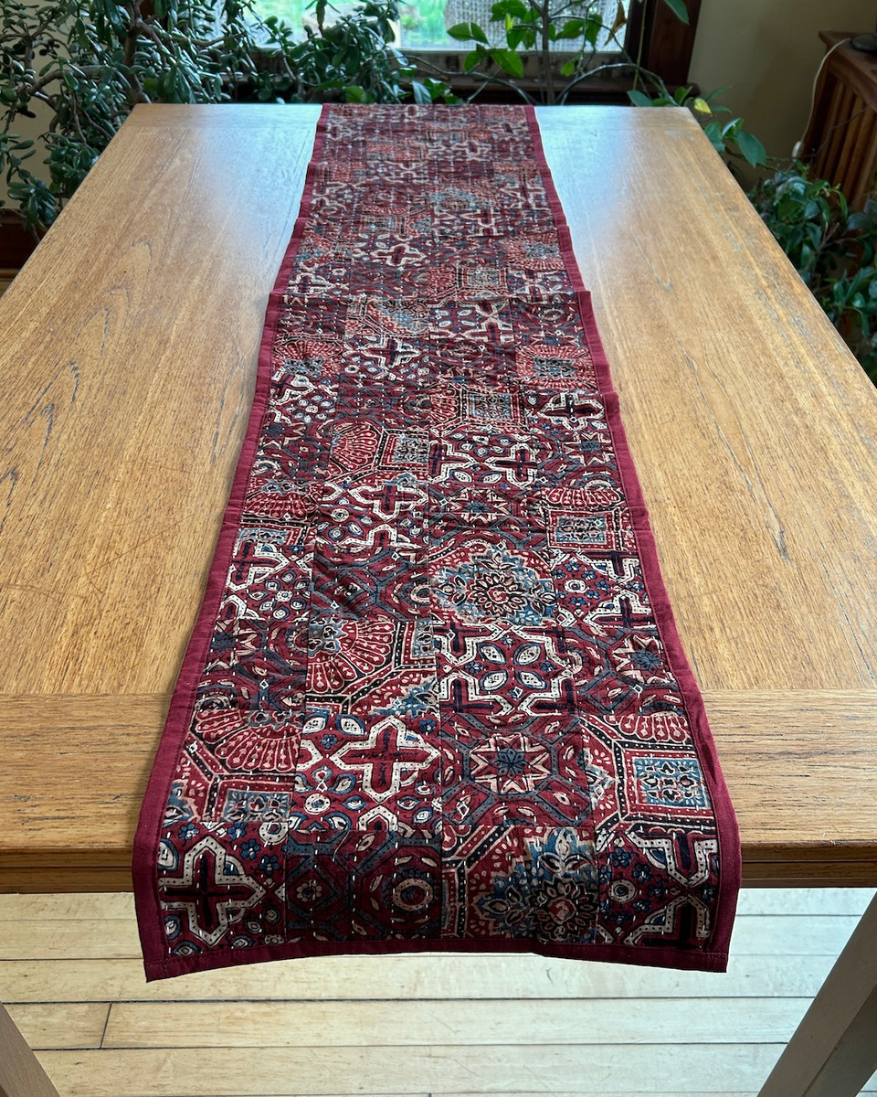 Handmade table runner of block printed fabrics. The fabrics are made with several wooden blocks using the traditional Ajarakh natural dye technique. The fabric is then sewn into a patchwork of various  patterns.  Colors: shades of brick red, cream, blue grey and black. This is reversible with the back solid block printed pattern in the same colors. There is a solid 1" brick red border sewn around.