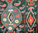 detail Suzani hand stitched wall hanging.  Suzanis are created by the once nomadic people who populated the ancient silk road of Uzbekistan. Suzanis were originally crafted as dowry pieces that a mother would help lovingly create for her daughter’s wedding day. It was believed that the patterns and colors in a suzani held talismanic powers of protection and these powers were magically transferred from the mother into the suzani through the visual impact of her embroidery work. From the studios of Sanjar Nazaroz,  a UNESCO Award of Excellence winner. This size makes a beautiful wall hanging. Shimmering forest green silk ground cloth. Embroidery colors: brick red, chalky teal green, cream, chalky pink, light indigo blue white and black.  There is a 1/2" black and tan woven band around the outside.