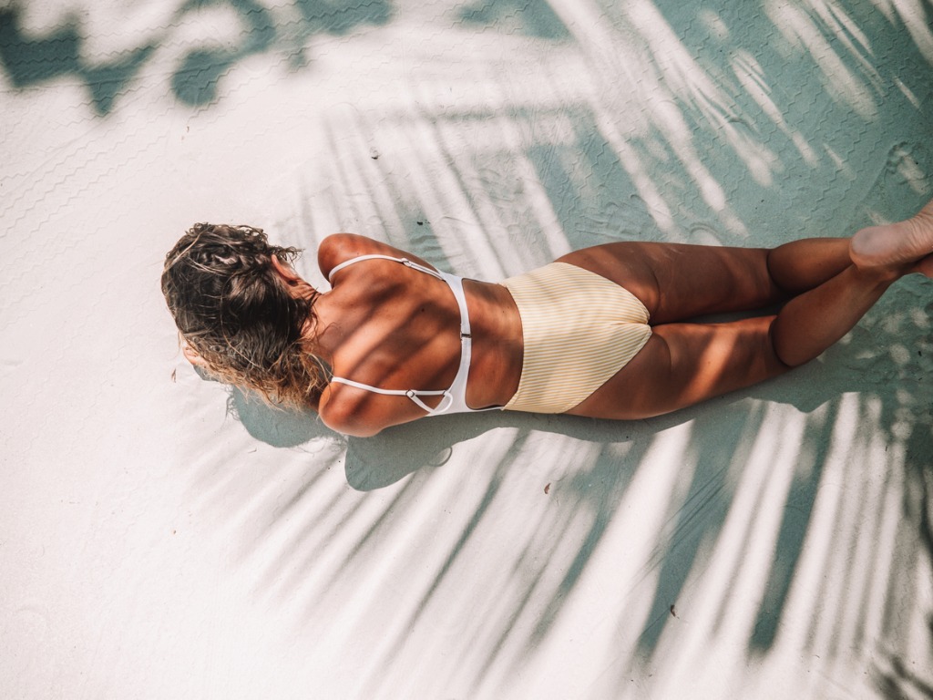 A stunning model lays on a sandy beach, basking in the sun's warm glow. The image showcases the model's beautifully bronzed skin after a flawless spray tan application. Playful shadows dance across the sand, adding depth and dimension to the scene.