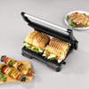 Electric grill with open lid and vegetable skewers, drip tray being removed by hand