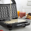 Black waffle maker open on kitchen counter, with waffles and fruit juice in background