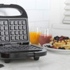 Black waffle maker open on kitchen counter with nearby orange juice and waffles