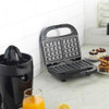 Kitchen scene with black waffle maker and juicer, plus plated waffles, juice and fruit.