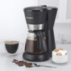 Black filter coffee maker with glass jug, next to cup of coffee, coffee beans and sugar bowl.