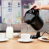 Black electric kettle pouring hot water into white teacup on kitchen table
