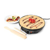 Electric crepe maker with cooked crepe, strawberries, chocolate sauce, and wooden utensils